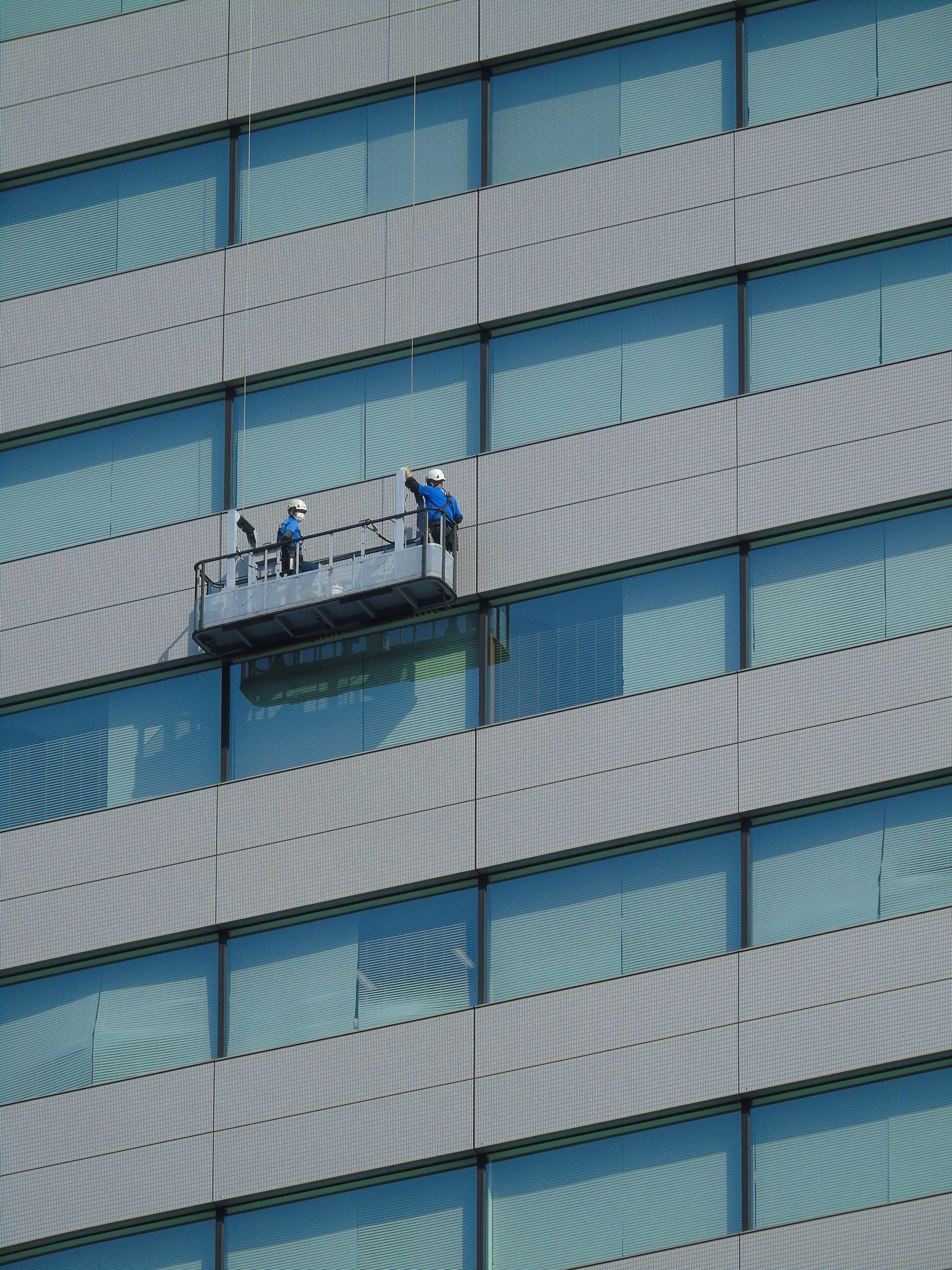 Two workers clean windows on a modern glass building facade using a suspended platform.